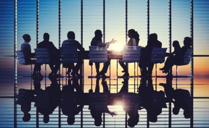 Men and women sitting around a board room table with sun shining through glass wall. Adobe